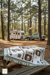 Quilt with nature-themed design on a wooden table in a forest setting with an RV and truck in the background.
