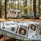Quilt with nature-themed design on a wooden table in a forest setting with an RV and truck in the background.
