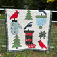 Quilt with winter-themed patterns including trees, birds, and a Christmas Stocking displayed outdoors.