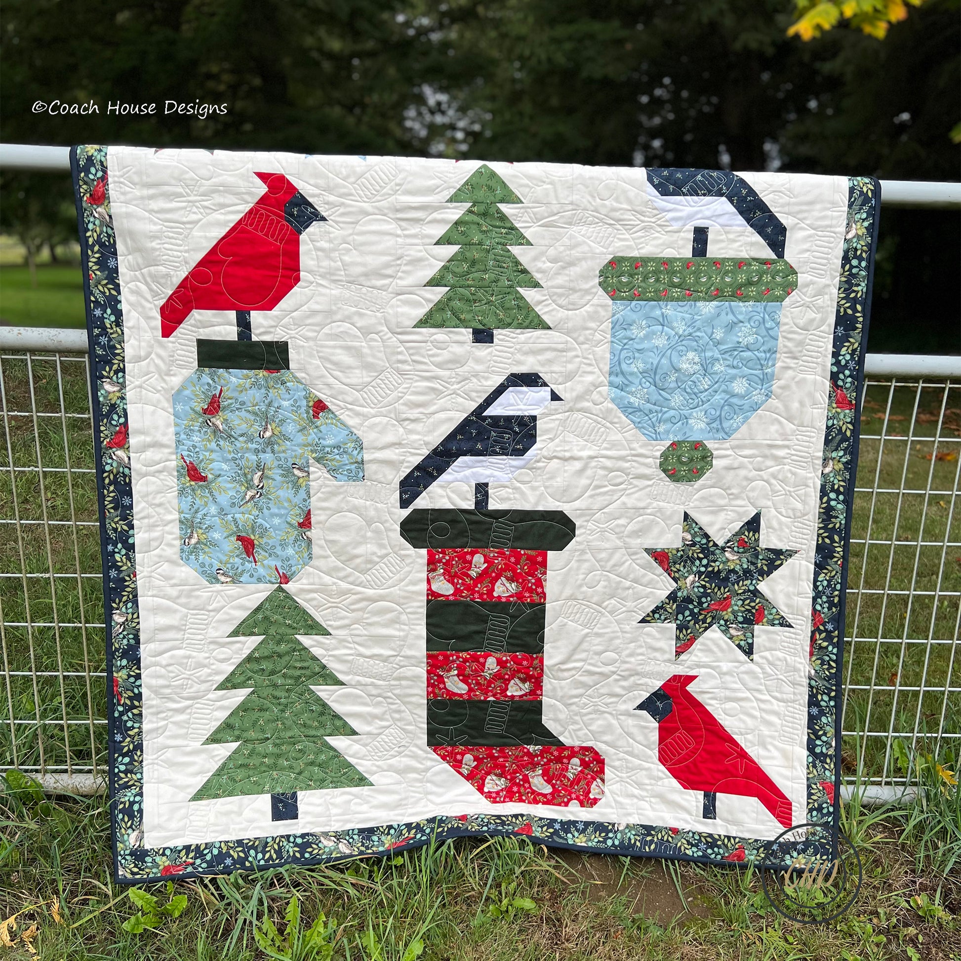 Quilt with winter-themed patterns including trees, birds, and a Christmas Stocking displayed outdoors.