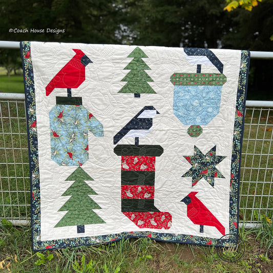 Quilt with winter-themed patterns including trees, birds, and a Christmas Stocking displayed outdoors.